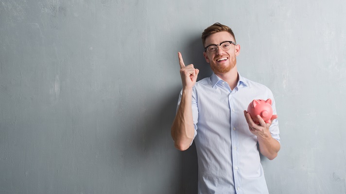 Young redhead man over grey grunge wall holding piggy bank surprised with an idea or question pointing finger with happy face, number one